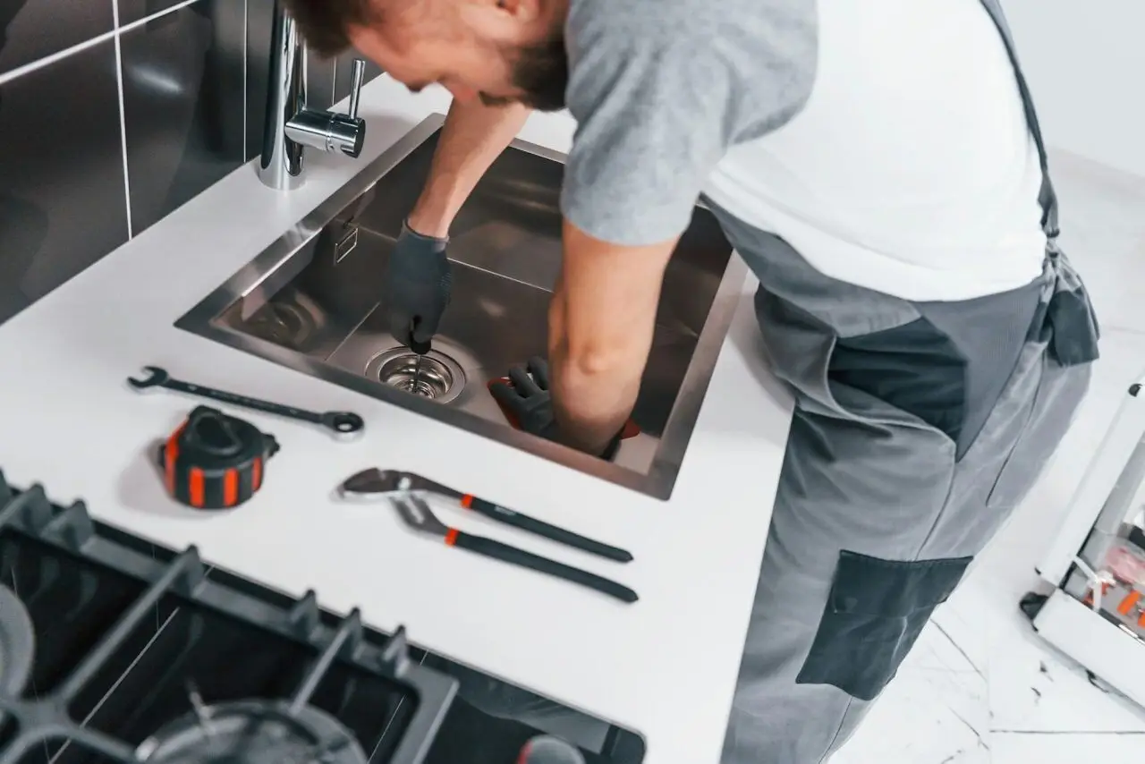 Young professional plumber in grey uniform repairing kitchen plumbing under the sink – PAGE Tradie Plumbing & Gas Engineers